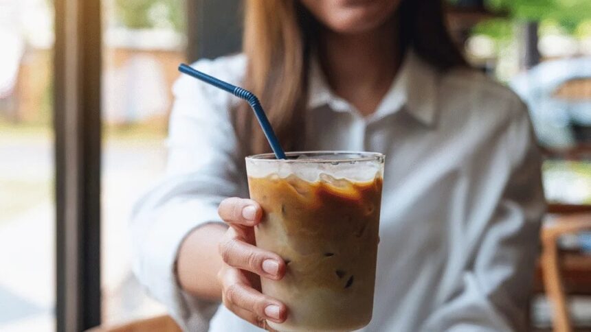 Mão segura copo de café gelado com canudo azul e camisa branca ao fundo