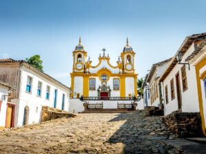 Igreja barroca amarela e branca ao fim de rua de paralelepípedos com casas coloniais