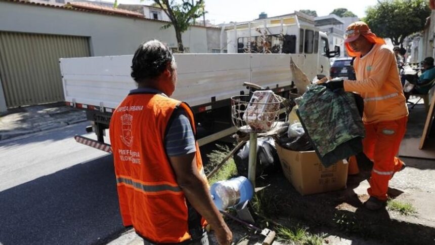 Dois agentes da Prefeitura de BH em uniformes laranja coletam lixo com caminhão ao fundo