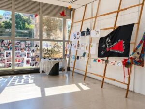 Exposição Mães de Maio em galeria: janelas com fotos, mesa e bandeira preta