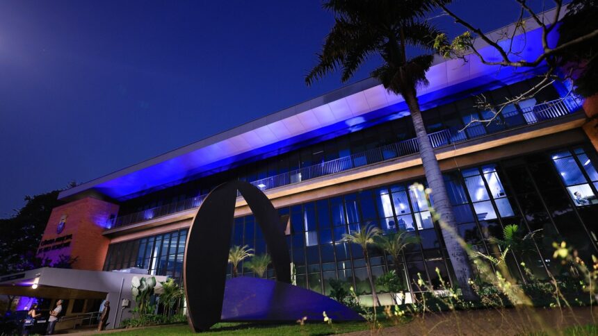 Fachada noturna da Câmara Municipal de BH, iluminada em azul, com palmeiras e escultura abstrata