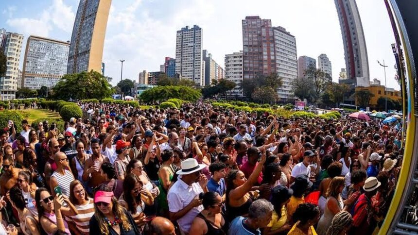 Multidão de pessoas em evento diurno ao ar livre, com edifícios urbanos e céu azul ao fundo