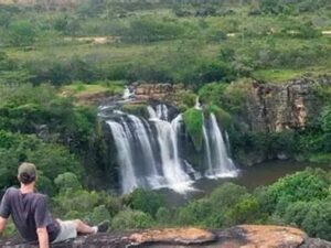Homem de boné e camiseta sentado em rocha, observando grande cachoeira em ambiente natural