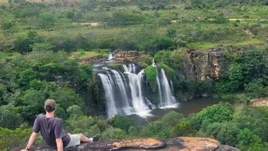 Homem de boné e camiseta sentado em rocha, observando grande cachoeira em ambiente natural