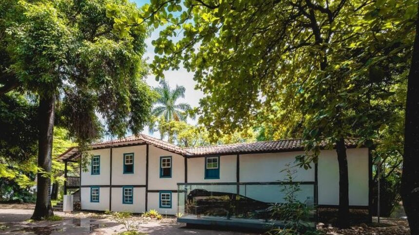 Fachada de um casarão branco e azul cercado por árvores frondosas sob o céu claro