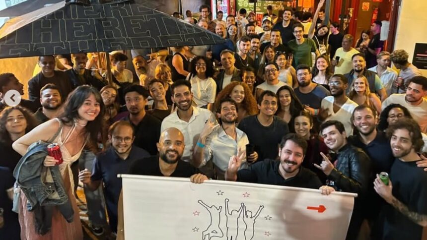 Grande grupo de pessoas sorrindo em evento noturno, com banner e guarda-chuva escrito HERE