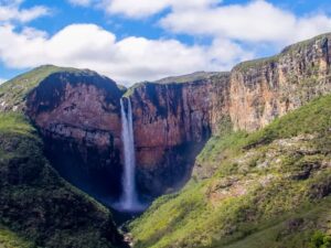 Cachoeira alta caindo entre paredões rochosos íngremes e vegetação verde sob céu azul com nuvens