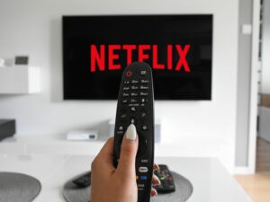 Mão segurando um controle remoto preto em frente a uma TV de tela plana exibindo o logo vermelho da Netflix em fundo preto, dentro de uma sala de estar com decoração clara e minimalista.