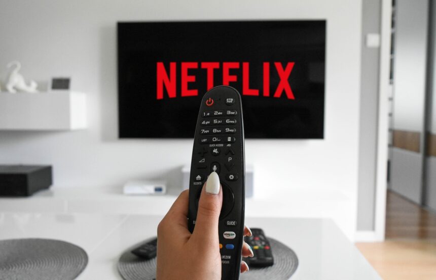 Mão segurando um controle remoto preto em frente a uma TV de tela plana exibindo o logo vermelho da Netflix em fundo preto, dentro de uma sala de estar com decoração clara e minimalista.