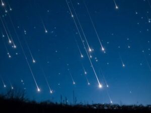 Céu noturno azul-escuro com múltiplas trilhas luminosas de meteoros caindo sobre uma paisagem escura.