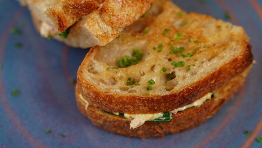 Sanduíche tostado, cortado ao meio, com recheio cremoso e cebolinha picada, em um prato azul