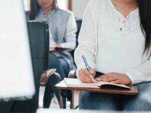 Mulher sentada, escrevendo em caderno com caneta azul, em sala de aula
