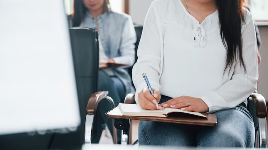 Mulher sentada, escrevendo em caderno com caneta azul, em sala de aula