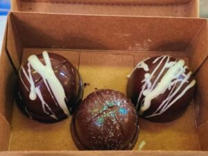 Três bombons de chocolate escuro em caixa de papelão, dois com cobertura branca, um brilhante