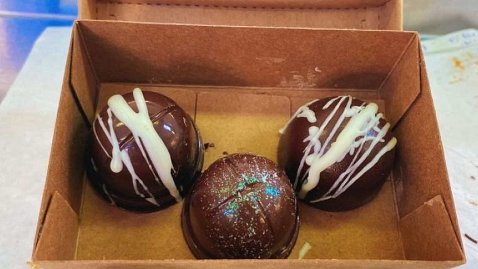 Três bombons de chocolate escuro em caixa de papelão, dois com cobertura branca, um brilhante