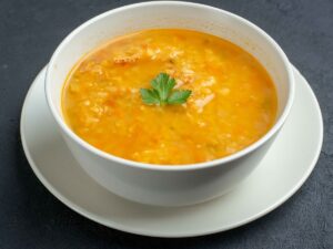 Tigela branca com sopa amarela, guarnecida com salsa, sobre pires branco e fundo escuro