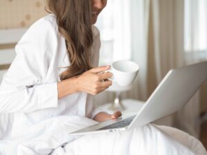 Mulher de cabelo castanho sorrindo, sentada na cama, usando laptop e segurando xícara branca