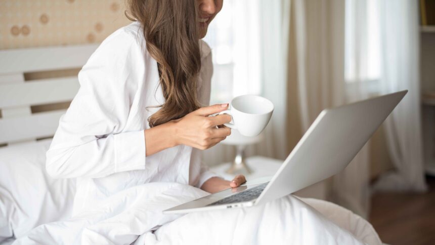Mulher de cabelo castanho sorrindo, sentada na cama, usando laptop e segurando xícara branca