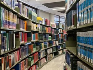 Corredor de biblioteca com prateleiras metálicas verdes repletas de livros coloridos, alguns com detalhes azuis.