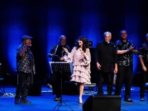 Cantora e seis músicos no palco em performance iluminada por luzes azuis