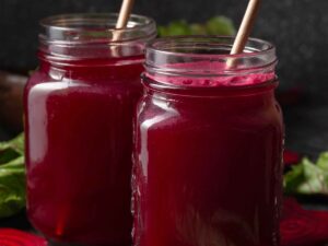 Dois copos de vidro com suco vermelho-escuro e canudos sobre fundo escuro com folhas e fatias de beterraba