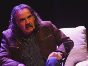 Humberto Martins, cabelo comprido e bigode, usando jaqueta marrom, sentado em cadeira com microfone