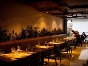 Interior de restaurante com mesas postas, vegetação na parede e funcionário organizando