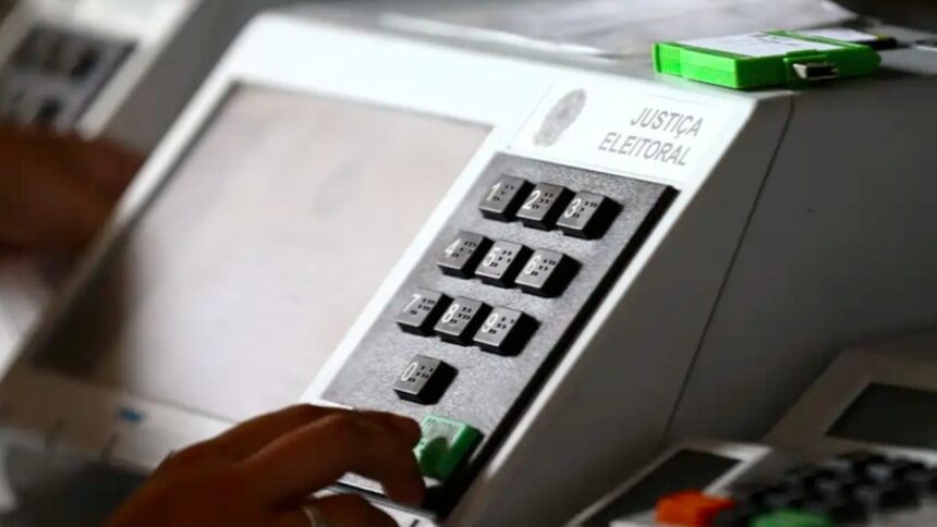 Mão pressiona botão verde de urna eletrônica com teclado numérico e braile. Inscrição 'JUSTIÇA ELEITORAL'