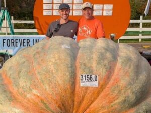 Dois homens sorrindo atrás de abóbora gigante com etiqueta 3156.0 RON WALLACE