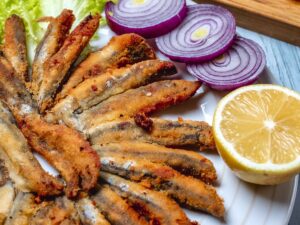 Pequenos peixes fritos em prato branco com alface, cebola roxa fatiada e meio limão