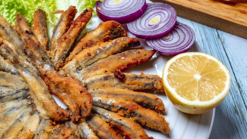 Pequenos peixes fritos em prato branco com alface, cebola roxa fatiada e meio limão