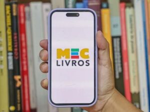 Mão segura smartphone exibindo o logo MEC LIVROS, com estante de livros ao fundo