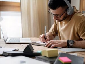 Homem com óculos e fones de ouvido escrevendo em caderno com laptop e materiais na mesa