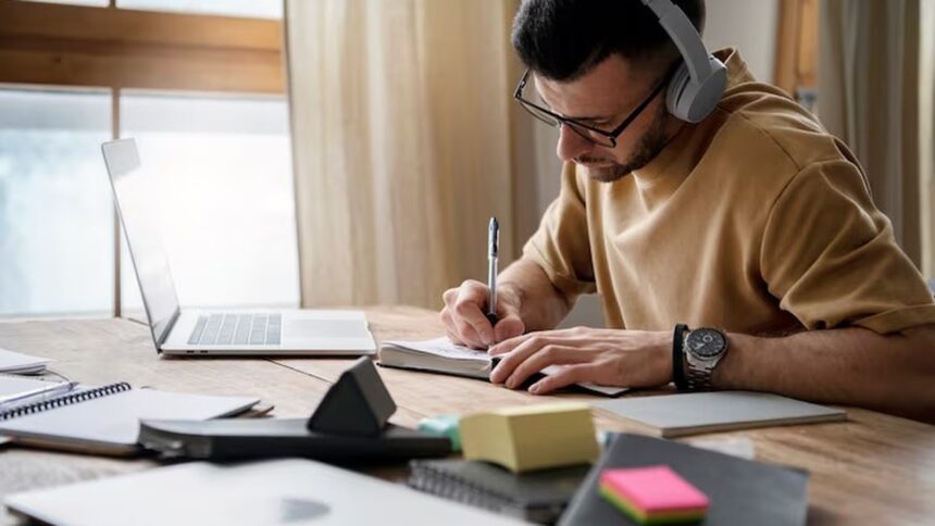 Homem com óculos e fones de ouvido escrevendo em caderno com laptop e materiais na mesa