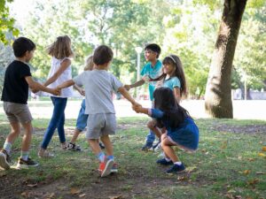 Sete crianças sorrindo, de mãos dadas em roda, brincando em parque com gramado