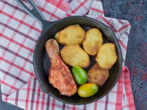 Coxa de frango assada, fatias de batata e frutas cítricas em frigideira de ferro sobre pano xadrez