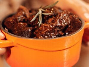 Mãos seguram panela laranja com carne escura em molho e um ramo de alecrim