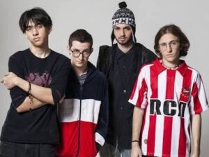 Quatro jovens homens, membros de banda, posam em estúdio, com gorro, óculos e camisa listrada
