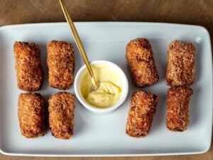 Prato branco com sete croquetes de frango fritos e tigela de molho com colher.