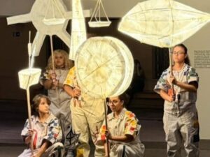 Cinco artistas seguram esculturas luminosas de peixe, relógio, balança e sol, no CCBB