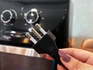 Mão feminina segurando um plugue de três pinos em frente a uma airfryer preta da marca Elgin