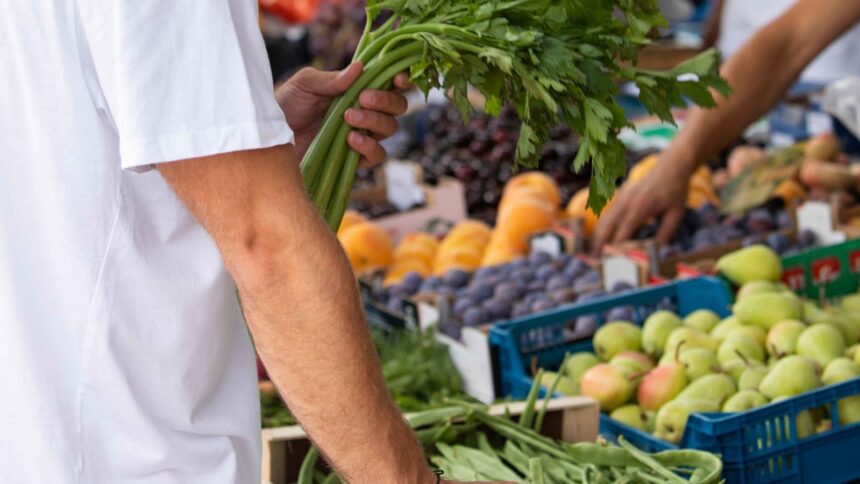 Pessoa segurando aipo fresco em feira de rua com barracas de frutas e legumes