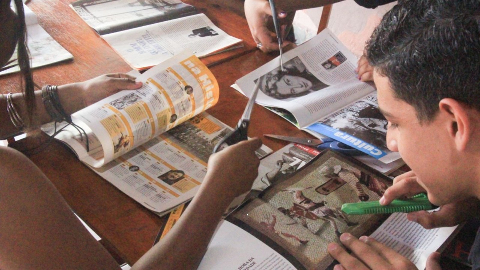 Mãos recortam revistas e jornais com tesouras e estilete em uma mesa de madeira