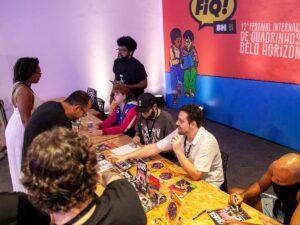 Autores autografam quadrinhos para fãs em mesa durante o Festival FIQ BH, com banner ao fundo