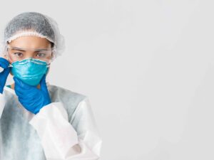 Profissional de saúde em EPI: touca, óculos, máscara azul e luvas, ajustando a máscara