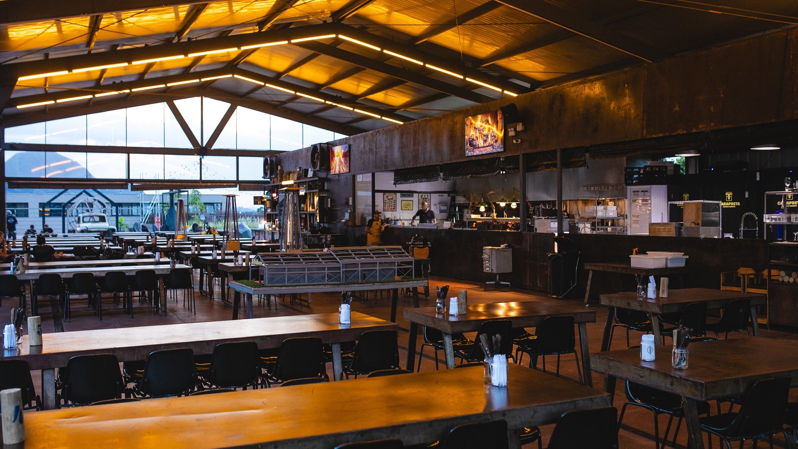 Interior de restaurante amplo com mesas de madeira, cadeiras, teto iluminado e cozinha aberta