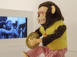 Macaquinho mecânico com colete amarelo batendo pratos. Ao lado, monitor exibe cena de filme