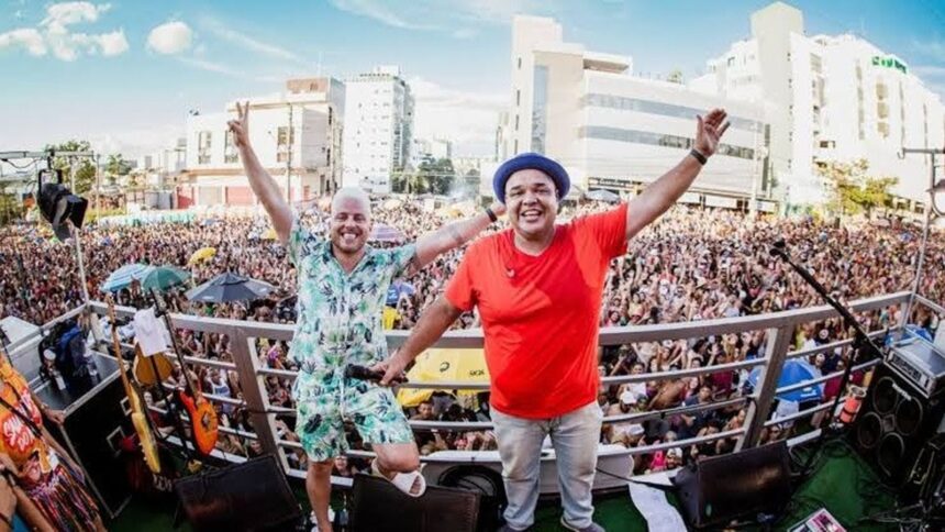 Dois homens sorrindo em palco, braços erguidos, para grande multidão em evento diurno