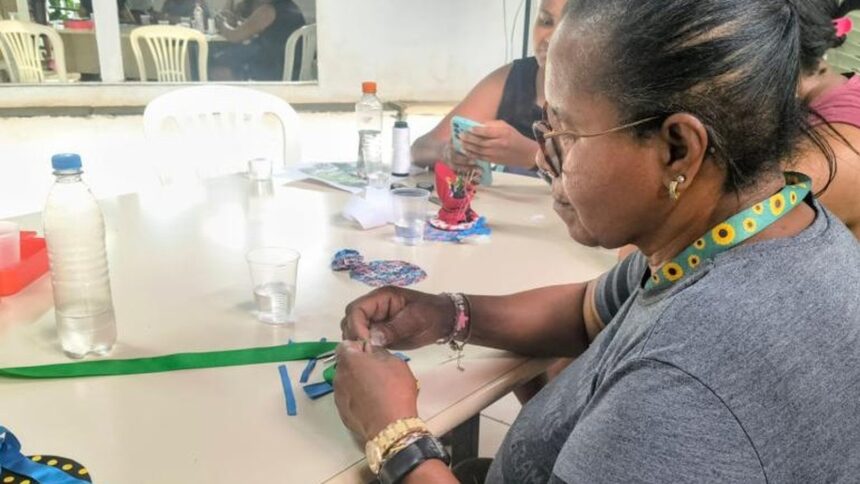 Mulher de óculos, sentada à mesa, fazendo artesanato com pedaços de tecido colorido