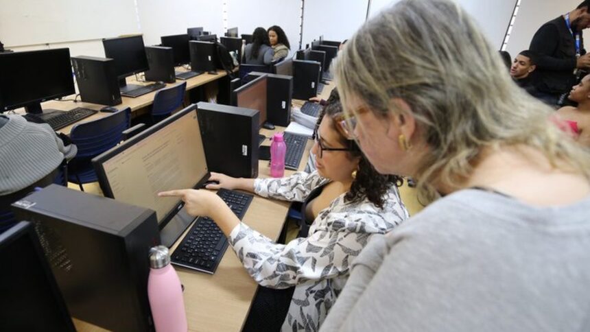 Duas mulheres em um laboratório de informática, uma aponta para a tela do computador